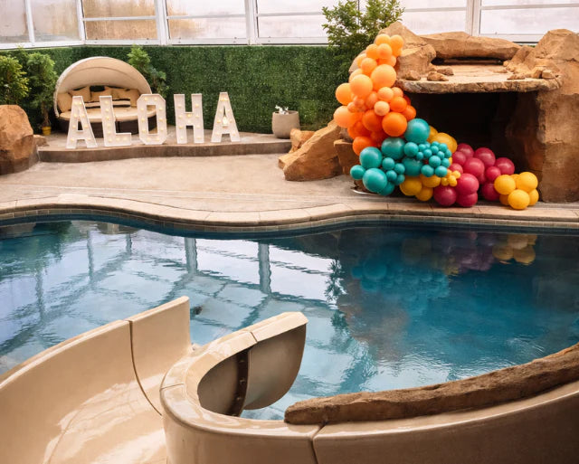 Pool area with colorful balloons and 'Aloha' letters near a water slide.