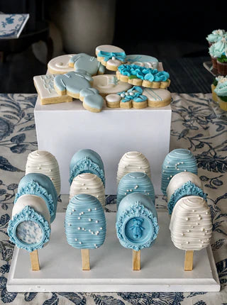 Decorative cookies on sticks and decorated cookies on a stand with a floral tablecloth background.
