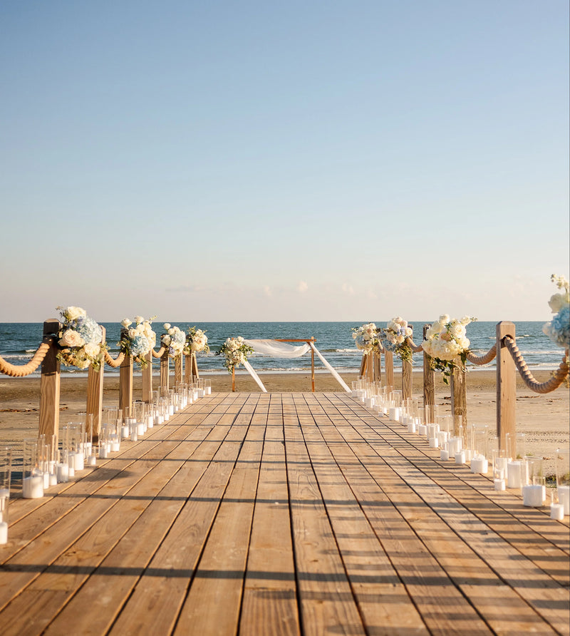 Decorated wooden walkway on a beach with floral arrangements and candles leading to the ocean.
