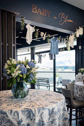 Display of baby clothes with 'Baby in Bloom' branding in a room with tables and chairs.