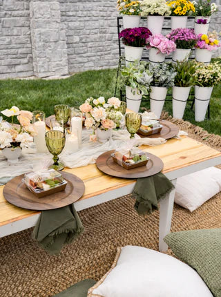 Outdoor dining setup with a wooden table, floral arrangements, and candles on a grassy area.