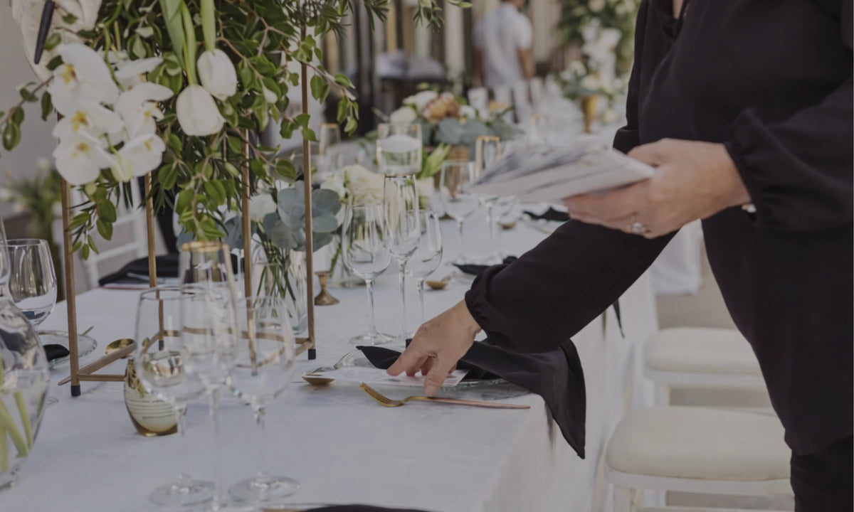 Person setting a table with white flowers and glasses at an event.