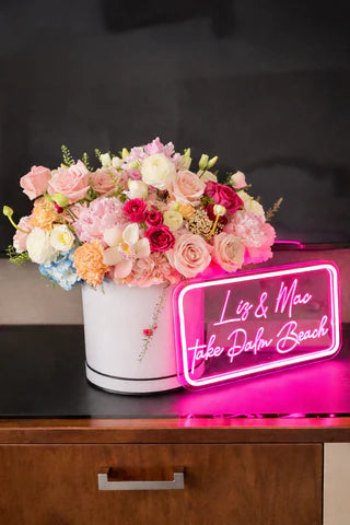 Neon sign with 'Liz & Mac' and 'Take Palm Beach' text next to a floral arrangement in a white box.